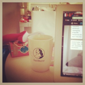A table featuring a box of blue kegel balls with a pink and white vibrator on top of it. In front of that is a coffee cup bearing the Good Vibrations logo beside a laptop, only the left third of the screen visible. 