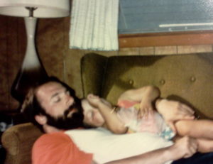 Bearded man asleep on a couch with little girl in Wonder Woman bathing suit asleep on his chest