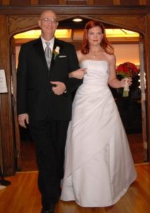 JoEllen in a strapless white dress with a long, full skirt holding a bouquet of red roses and arm in arm with a bald man in a black suit