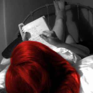 Woman lying on a bed with her legs up on the wrought iron headboard as she writes in a notebook. The top of her head is facing the camera. The photo is black and white and her hair is bright red. 