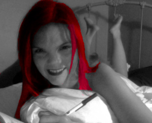 Woman on her stomach on a bed writing in a notebook, her feet and the wrought-iron headboard visible in the background. The photo is black and white but her hair is bright red.