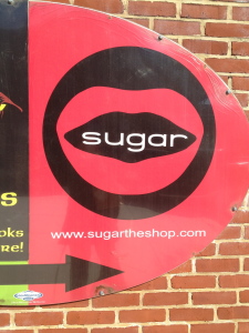 This Way To Sugar! A red sign featuring a black circle with red lips and the word "sugar" in the middle. Below is a black arrow pointing toward the store
