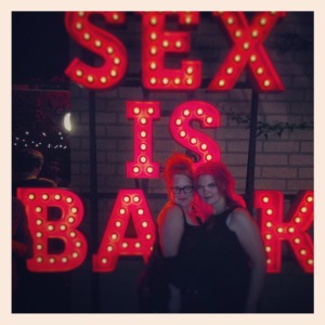 Two women standing in front of a large red neon sign reading "Sex is Back"