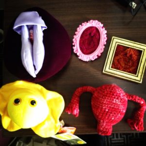 Table holding a vulva puppet, a crocheted uterus, 2 knit vulvas and a plush model of the herpes virus