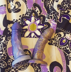 Two Überrime dildos--one purple and one purple and gold-- on a beige dress with a purple paisley design. 